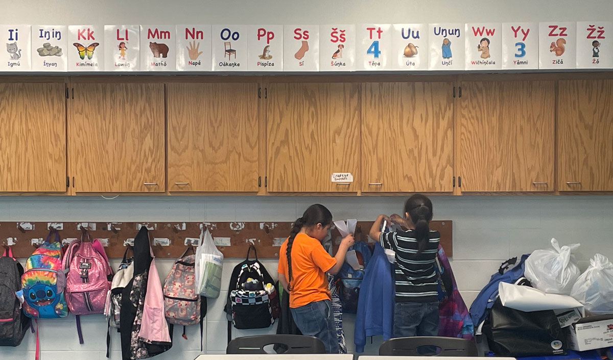 Two Native American students hang their materials on a coat rack in a classroom at the Red Cloud Indian School, where letters of the Lakota alphabet are displayed on the wall above. South Dakota News Watch photo by Bart Pfankuch